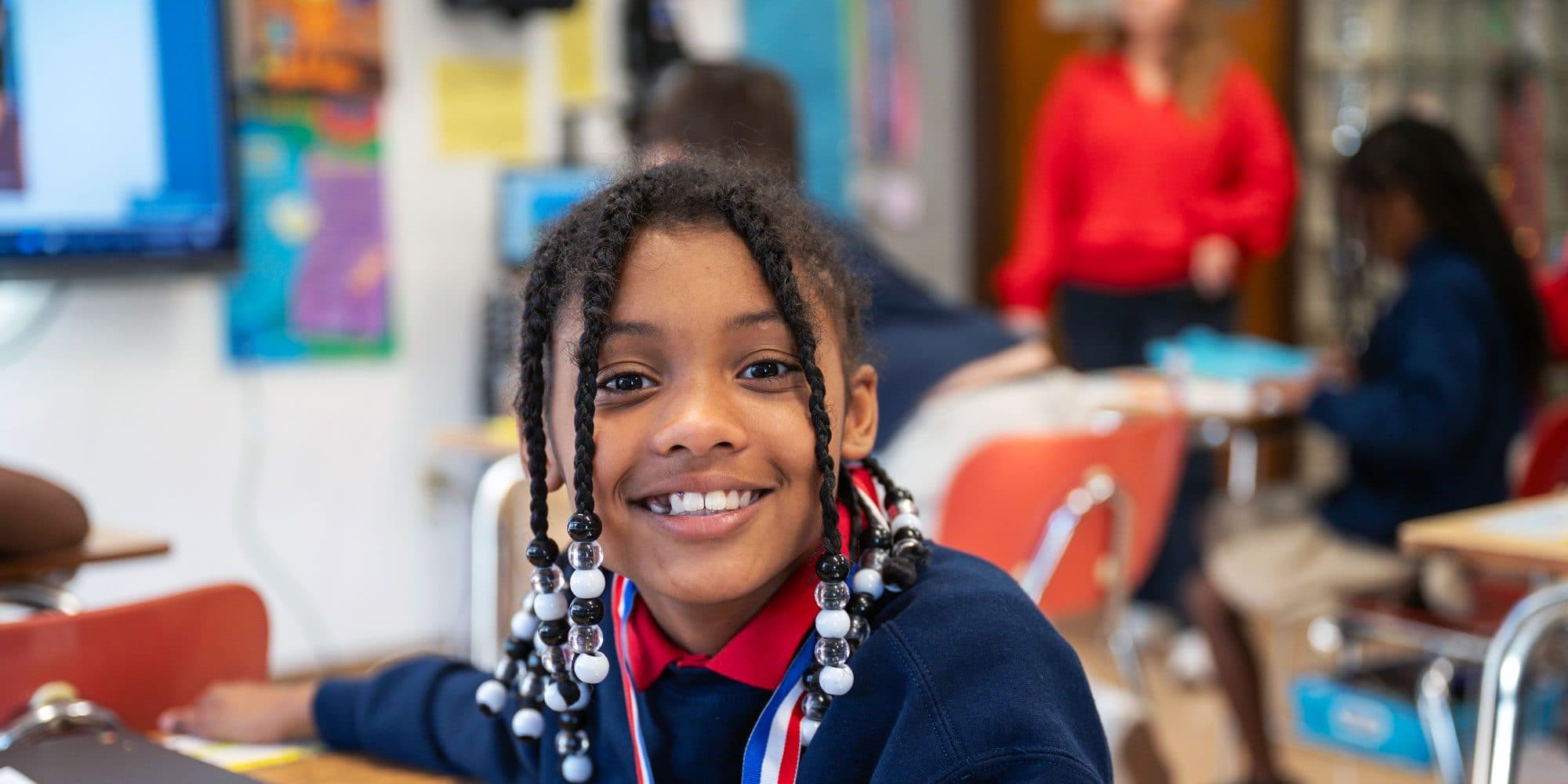 Smiling student with braided hair and beads at a classroom desk.