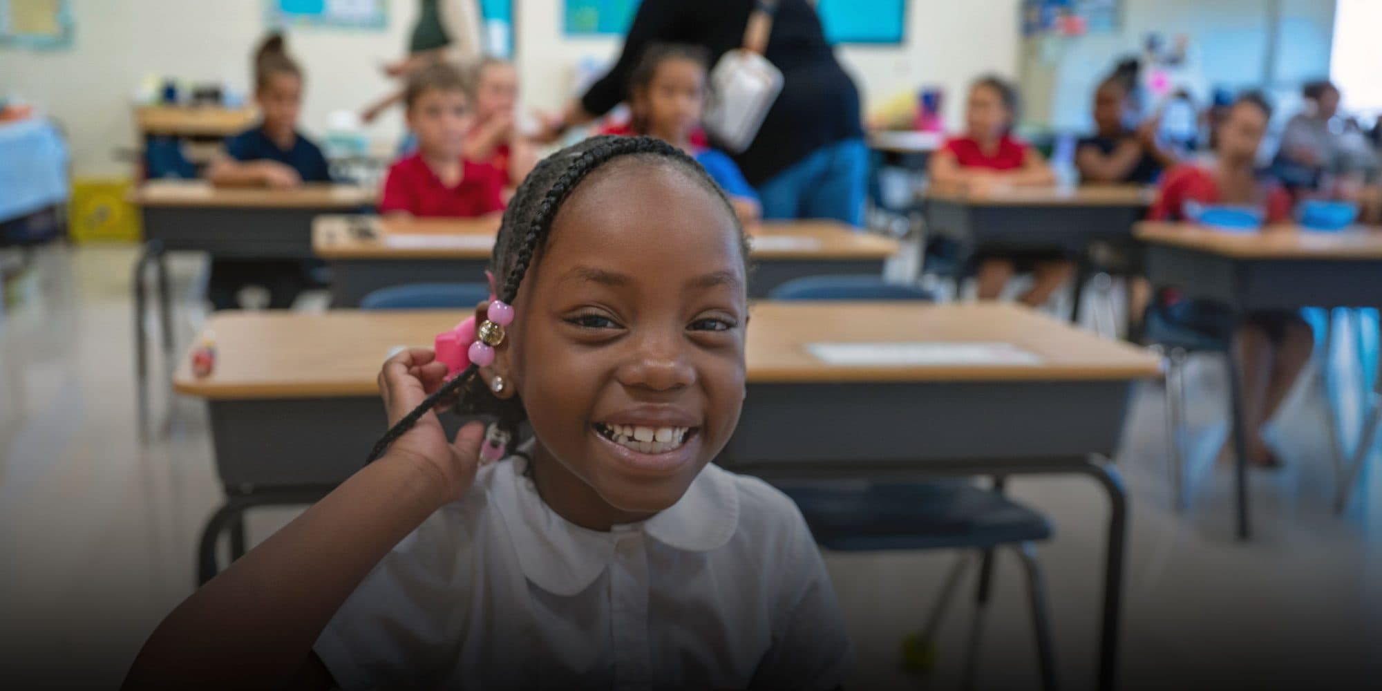 Smiling Foundation Academy student with braided hair and pink beads at a classroom desk.